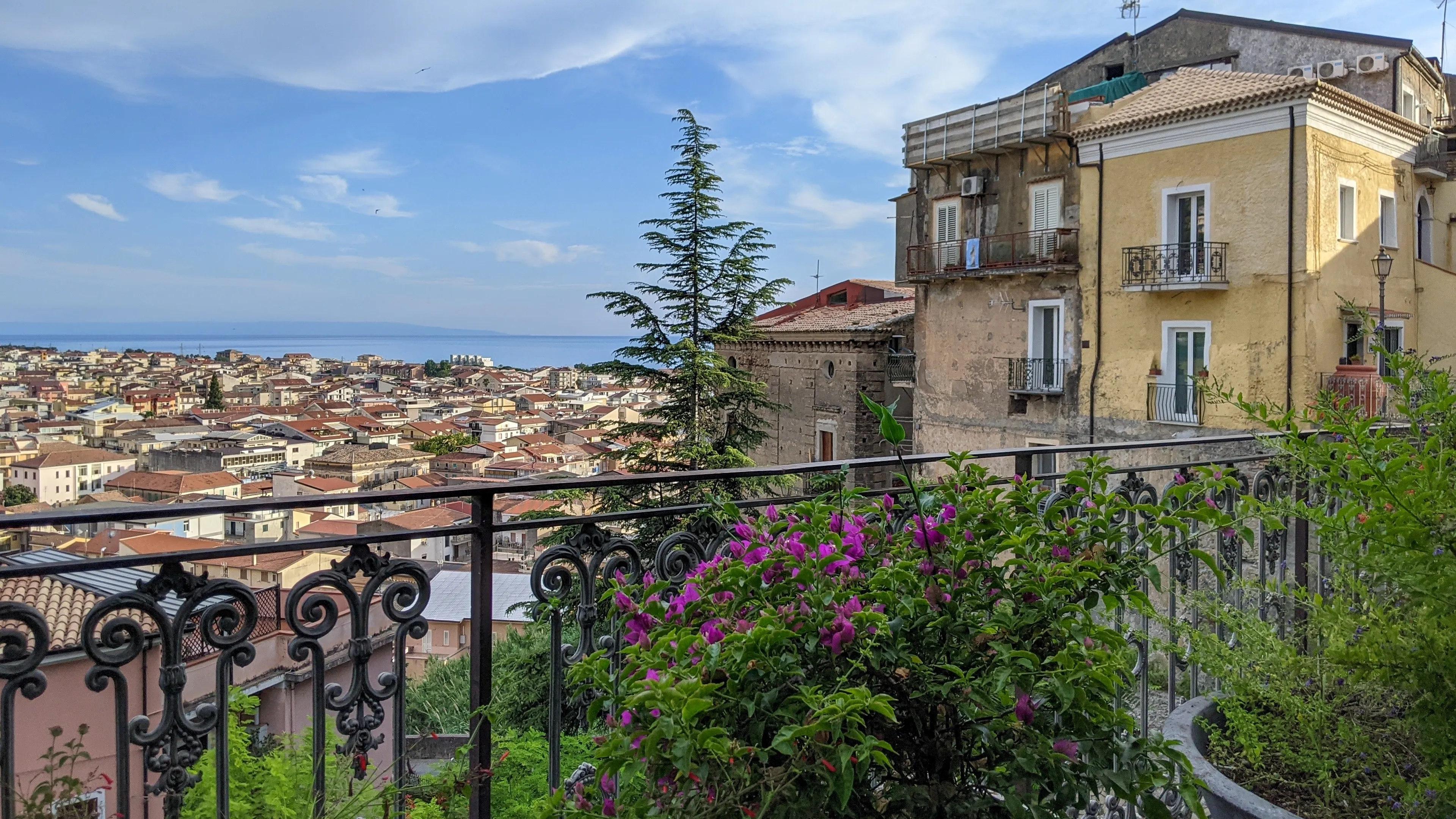 Amantea, Cosenza, Italy - July 4, 2021:Scenic overlook of Mediterranean town with terracotta roofs, ornamental iron balcony railing, flowering plants and cypress tree