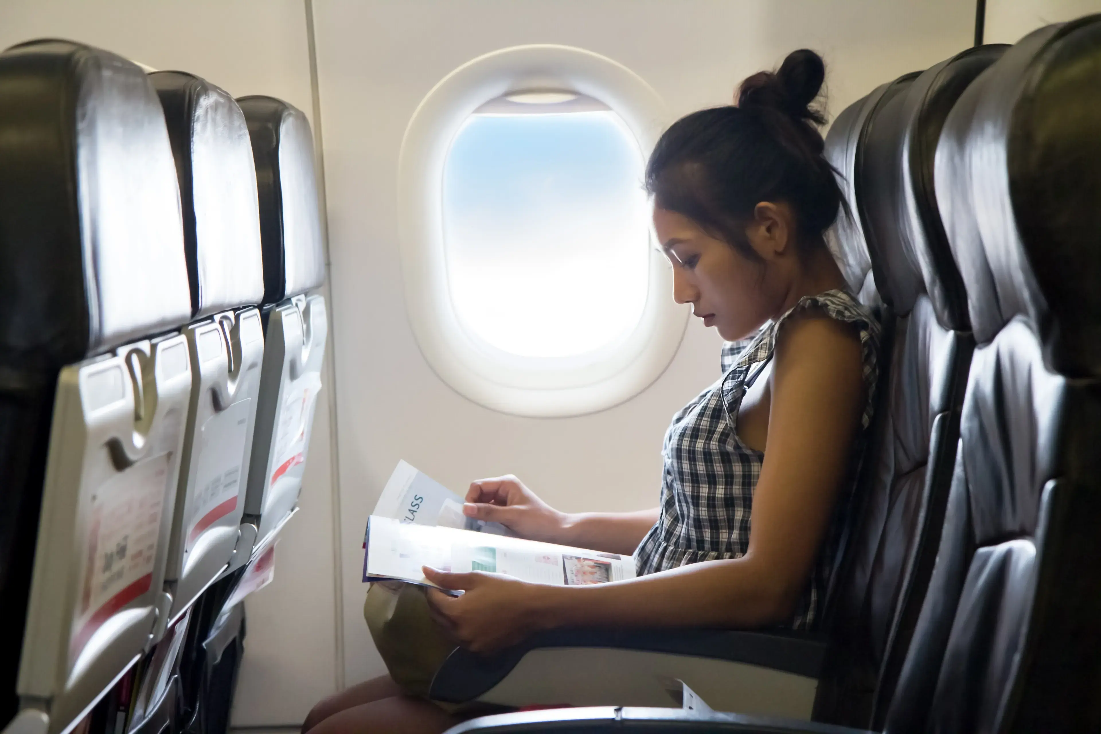 Young woman travels in a chair of the airplane. Passengers reads a magazine in the window of the aircraft. Enjoy - entertainment on board of flying air plane. Young woman travels in a chair of the airplane. Passengers reads a magazine in the window of the aircraft. Enjoy - entertainment on board of flying air plane.