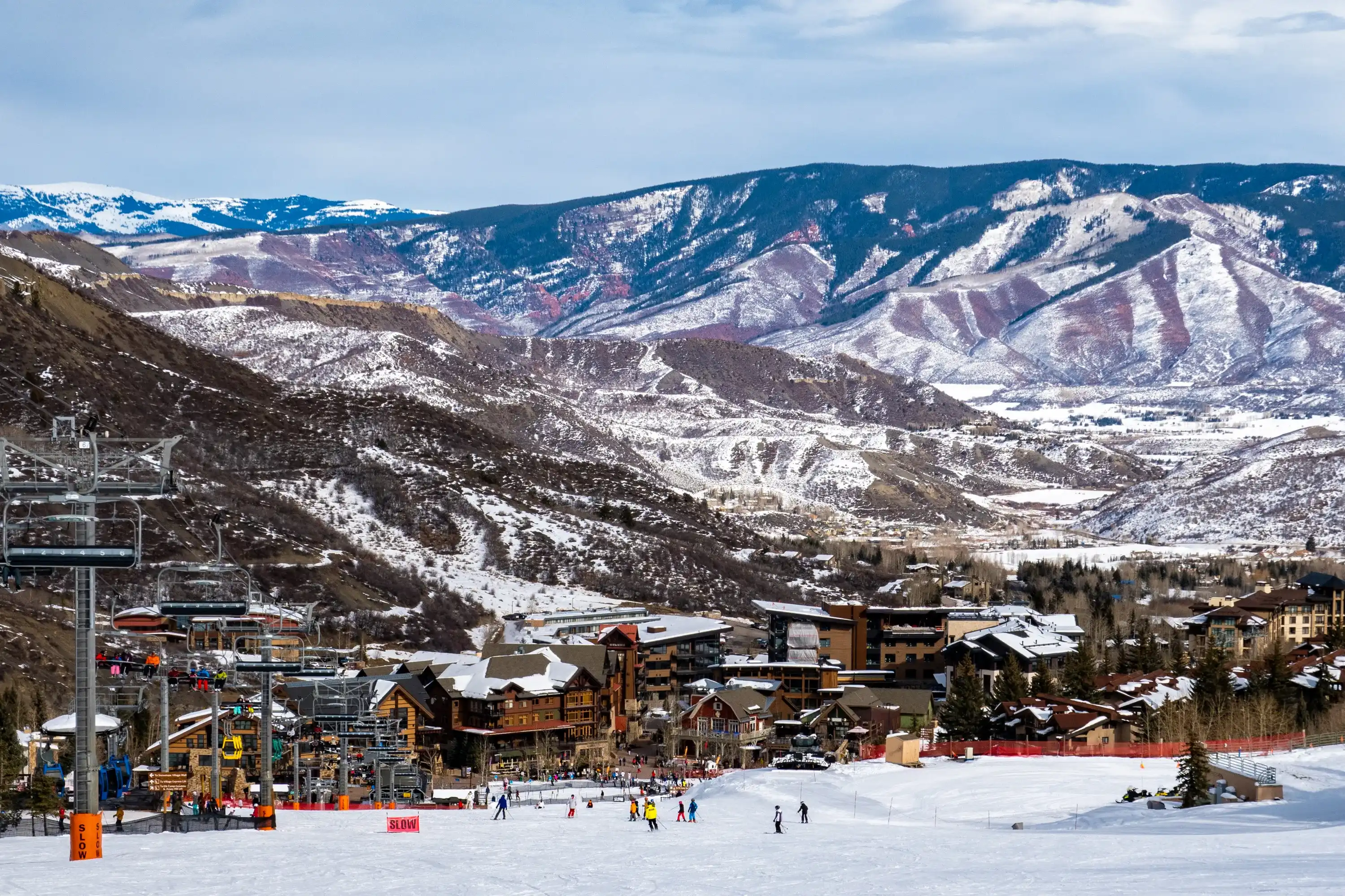 Panoramic view of Snowmass Village, with skiers skiing at the Aspen Snowmass ski resort in the foreground and the Rocky Mountains of Colorado in the background, on a partly cloudy winter day. Panoramic view of Snowmass Village, with skiers skiing at the Aspen Snowmass ski resort in the foreground and the Rocky Mountains of Colorado in the background, on a partly cloudy winter day.