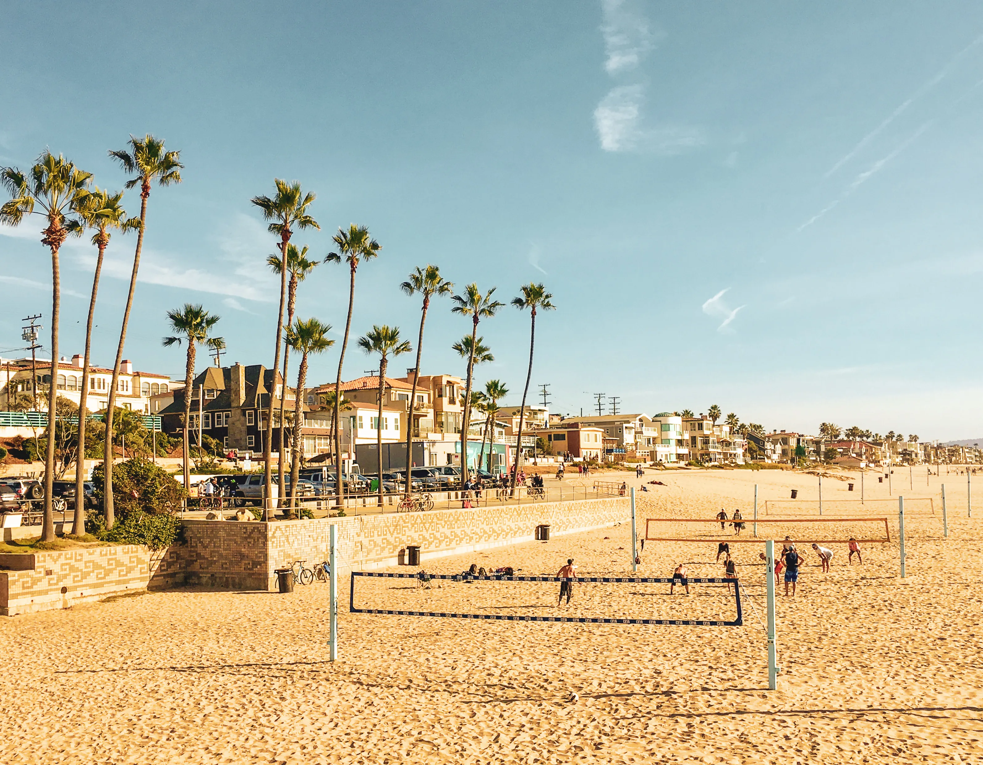 Tall palm trees, parking lot, beachfront homes on the Strand, and people playing beach volleyball in Manhattan Beach, Los Angeles, California. Travel, urban living, active lifestyle and sports concept