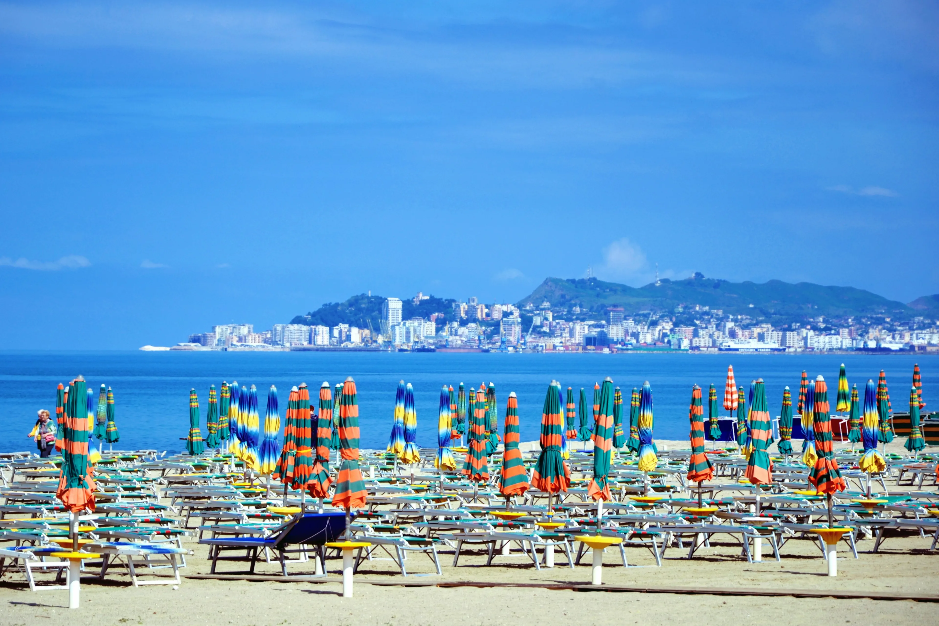 Closed umbrellas and empty chaise lounges at the sandy beach. Beach chairs, beds and umbrellas on the Albanian beach before summer holiday season. Golem, Durres, Albania. Durres city in a distance
