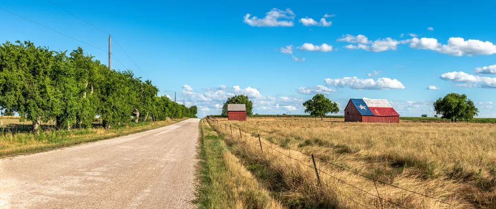 An abandoned old barn in Rural Texas with the state flag painted on its roof sits in a farmland community framed by fields and a dirt road. 