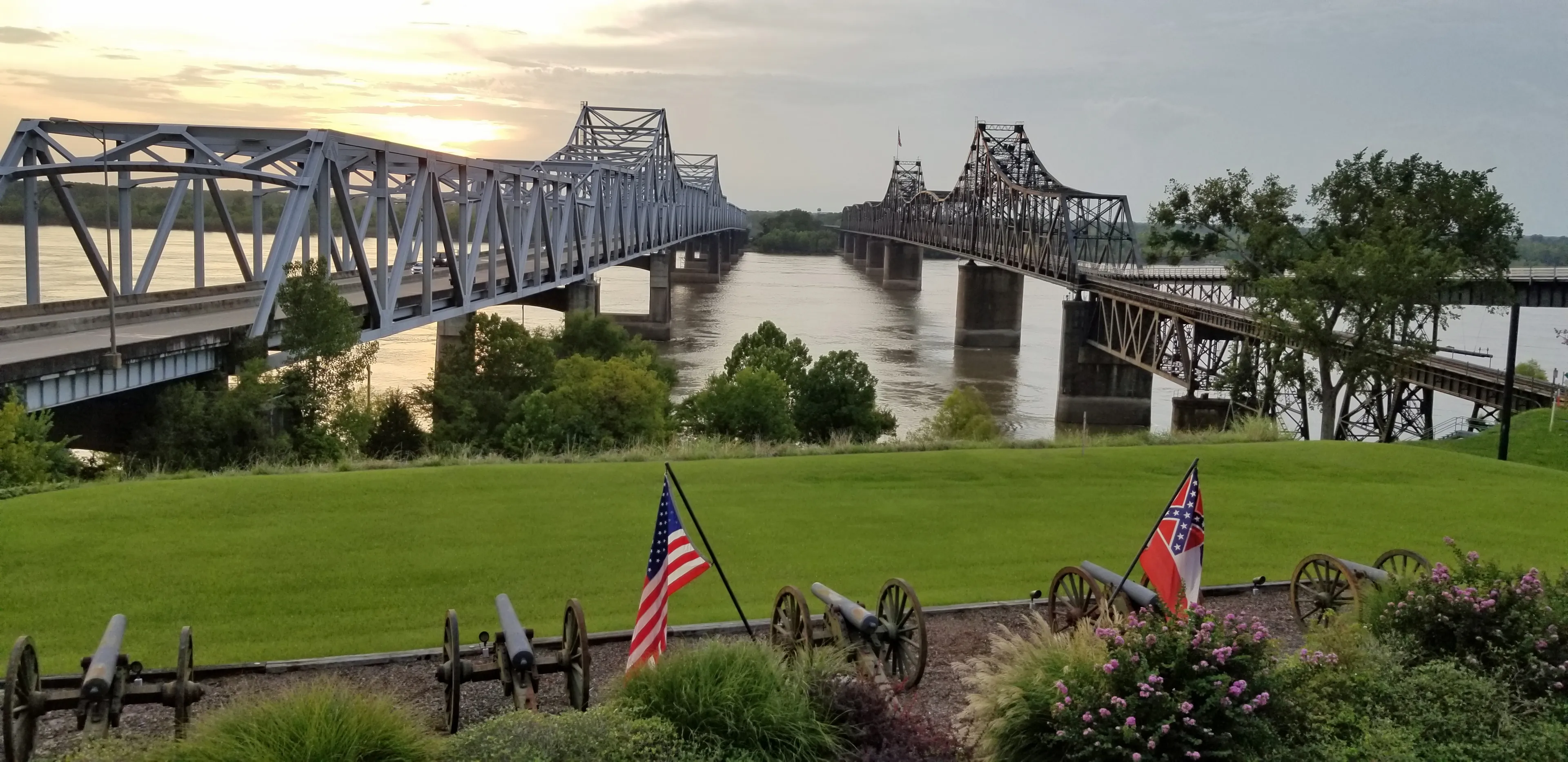 Two bridges crossing the Mississippi River contrasting old and new, railroad and highway, and train and road in Vicksburg, Mississippi with the United States and State of Mississippi flag flying.