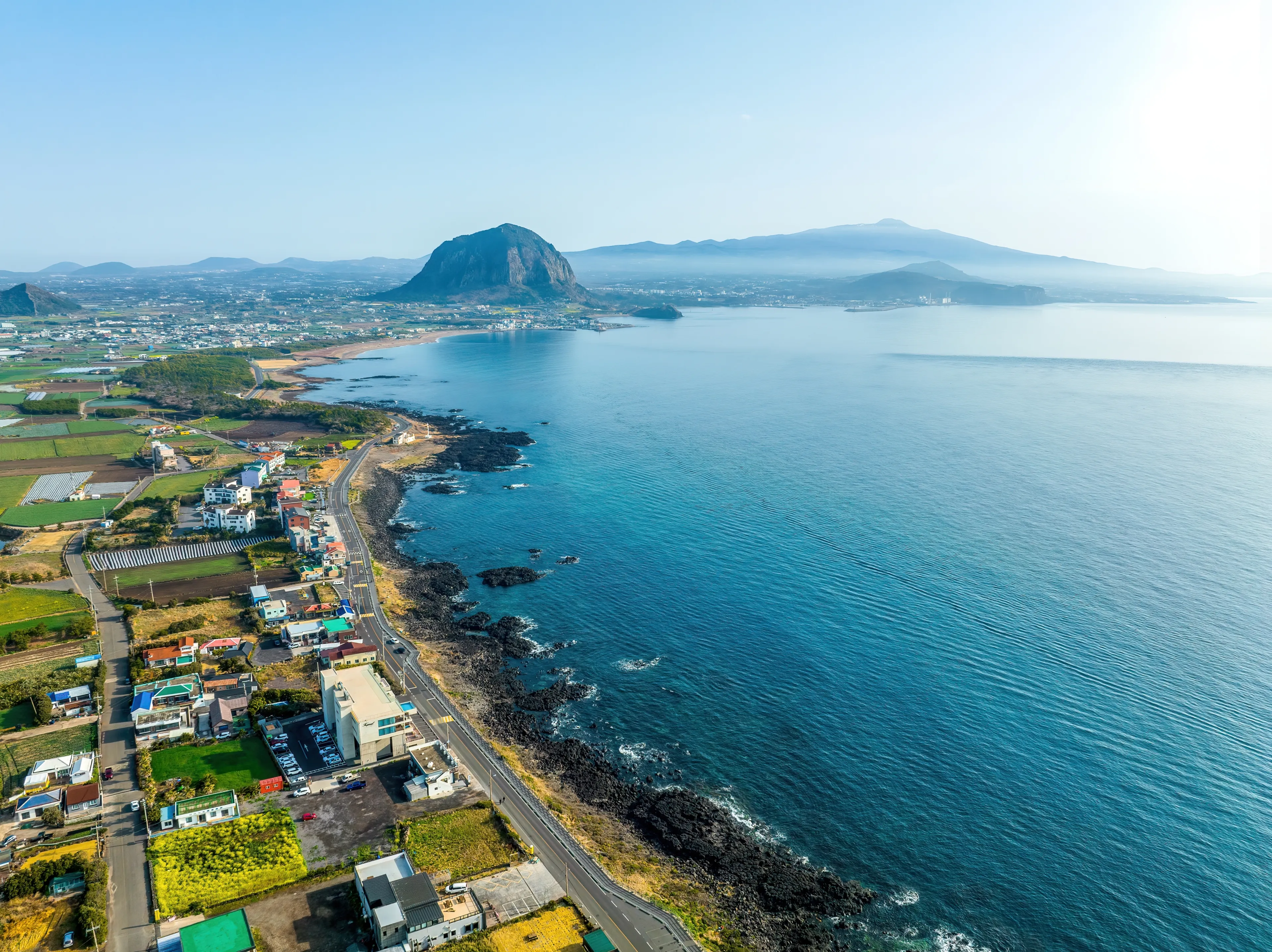 Daejeong-eup, Seogwipo-si, Jeju-do, South Korea - March 16, 2022: Aerial and morning view of road and houses of a village near Sagye Beach against Sanbangsan and Hallasan Mountain in the backgroud 